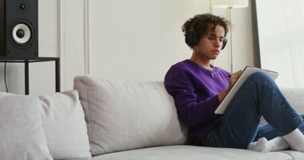 Handsome guy in headphones makes notes or draws in notebook sitting on sofa — Stock Video