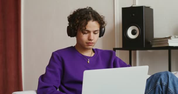 Knappe vent in een koptelefoon gebruikt een laptop zittend op een bank in een modern interieur — Stockvideo