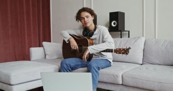 En kille med gitarr i händerna sitter på en soffa och ler mot kameran — Stockvideo