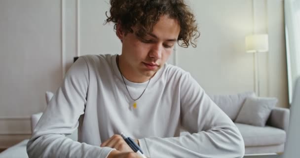 Όμορφος άνθρωπος κρατά σημειώσεις σε ένα σημειωματάριο, ενώ κάθεται μπροστά από ανοιχτό φορητό υπολογιστή — Αρχείο Βίντεο