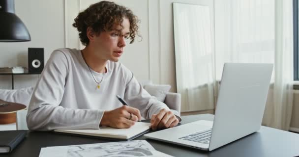 Hombre guapo hace notas en un cuaderno mientras está sentado frente a la computadora portátil abierta — Vídeos de Stock
