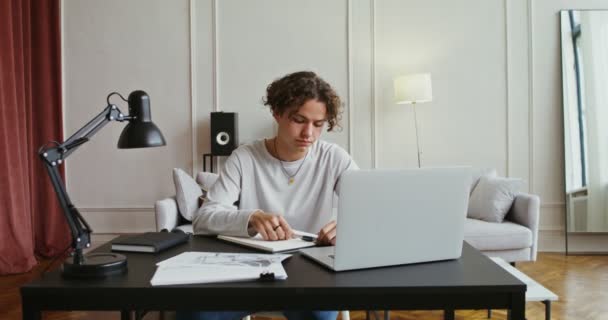 Knappe man maakt aantekeningen in een notebook terwijl hij voor een open laptop zit — Stockvideo