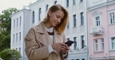 Genç bir kadın cep telefonuna bakıyor ve gülümsüyor, cadde boyunca yürüyor.