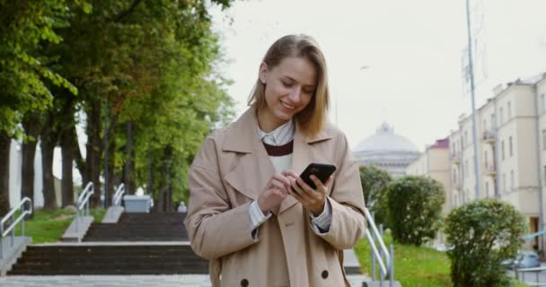 Молодая женщина смотрит в мобильный телефон и улыбается, идя по городской улице — стоковое видео