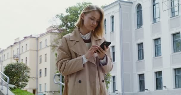 Genç bir kadın cep telefonuna bakıyor ve gülümsüyor, cadde boyunca yürüyor. — Stok video