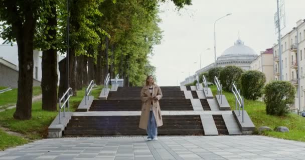 Una joven mira hacia el móvil y sonríe, caminando por la calle de la ciudad — Vídeos de Stock