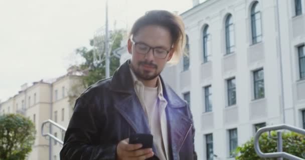 Ένας νεαρός άνδρας χρησιμοποιεί το τηλέφωνο ενώ περπατά στο δρόμο της φθινοπωρινής πόλης — Αρχείο Βίντεο