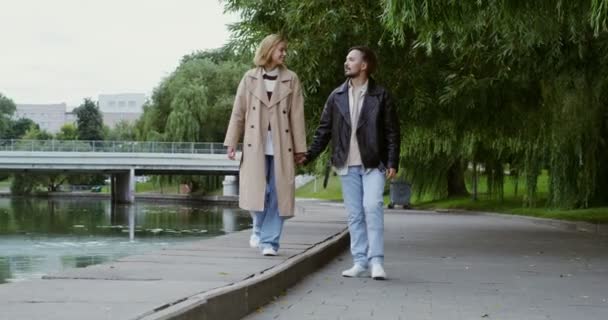 Casal jovem estão andando ao longo aterro ao longo do rio, de mãos dadas e falando — Vídeo de Stock
