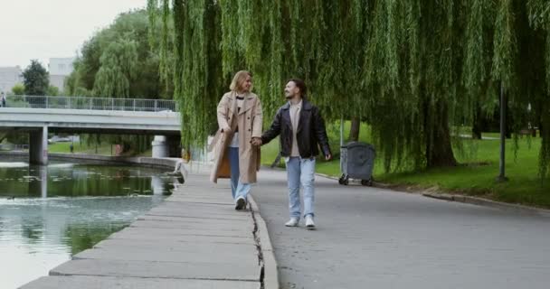 Fiatal pár sétál a parton a folyó mentén, fogják egymás kezét és beszélgetnek. — Stock videók