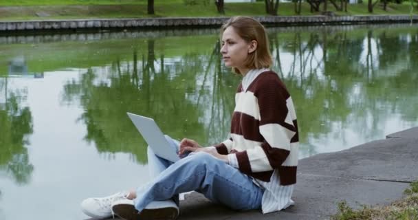 En ung flicka använder en bärbar dator som sitter på trottoaren på banvallen — Stockvideo