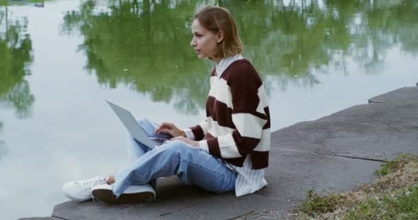 Uma jovem usa um laptop sentado na calçada no dique — Vídeo de Stock