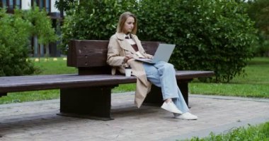 Genç bir kadın dizüstü bilgisayarda çalışıyor, bankta oturuyor ve kahve içiyor.