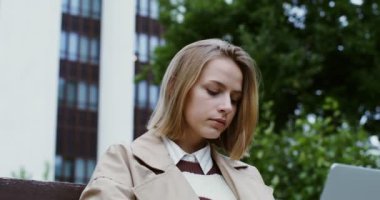 Genç bir kadın dizüstü bilgisayarda çalışıyor, bankta oturuyor ve kahve içiyor.