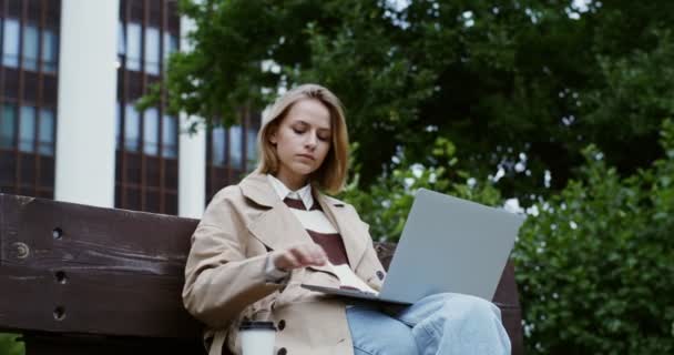 Jovem trabalha em um laptop, sentado em um banco de parque e bebendo café — Vídeo de Stock