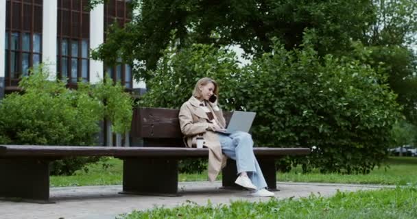 若い女性はラップトップで動作し、公園のベンチに座って携帯電話で話す — ストック動画