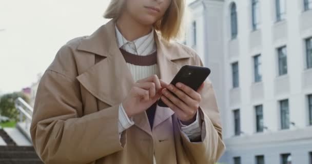 Una joven mira hacia el móvil y sonríe, caminando por la calle de la ciudad — Vídeo de stock