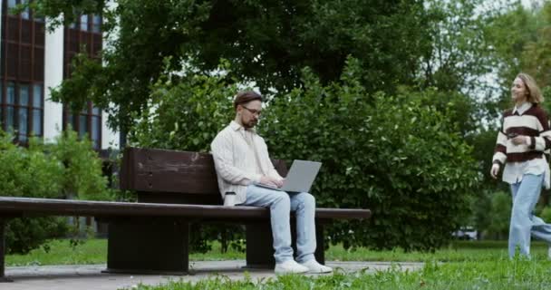 Una mujer se acerca a un tipo que trabaja en el portátil sentado en el banco y lo saluda — Vídeos de Stock