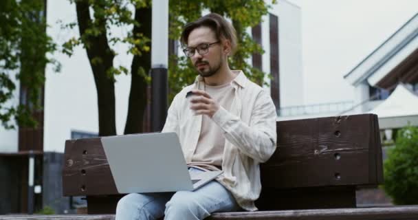 Een jonge man werkt op een laptop en drinkt koffie terwijl hij op een bankje in het park zit — Stockvideo