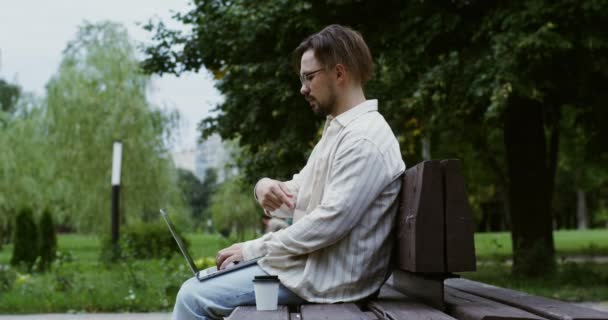 Een jongeman werkt op een laptop, praat via videogesprekken, zit op een bankje in het park — Stockvideo