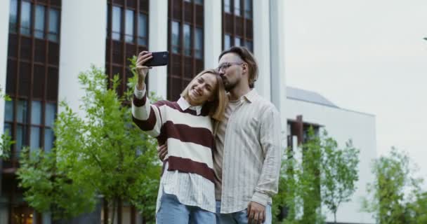 Fiatal férfi és nő mosolyognak és ölelkeznek, szelfit csinálnak egy mobilon a parkban. — Stock videók
