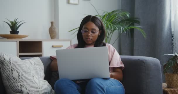 アフリカ系アメリカ人の女性は現代的なインテリアのソファに座ってラップトップを使用しています — ストック動画