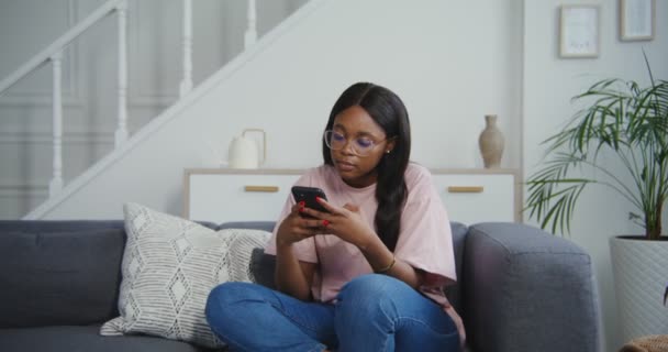 Afrikansk amerikansk kvinna ler när hon använder telefonen medan hon sitter i soffan — Stockvideo