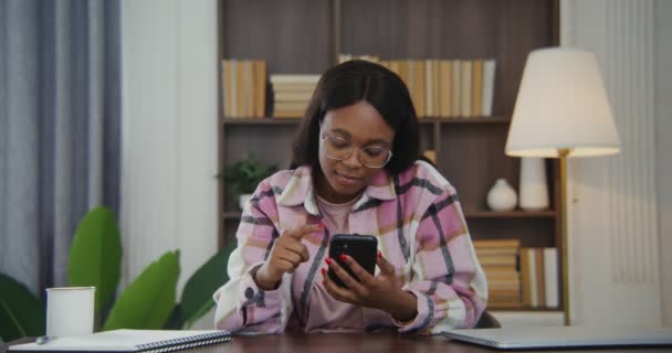 Afrikaans amerikaanse vrouw gebruikt telefoon terwijl zitten aan een bureau in een modern interieur — Stockvideo