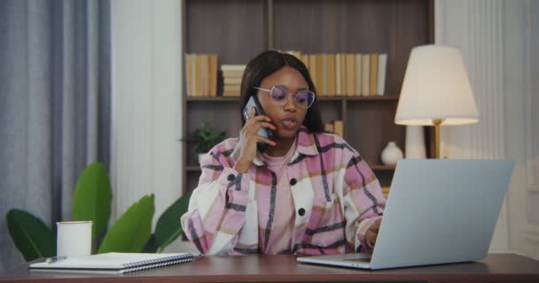 Afrikansk-amerikansk kvinne som snakker i mobiltelefon mens hun jobber på en bærbar datamaskin – stockvideo