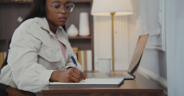 Африканская американка работает над ноутбуком, делая заметки в блокноте, сидя за столом — стоковое видео