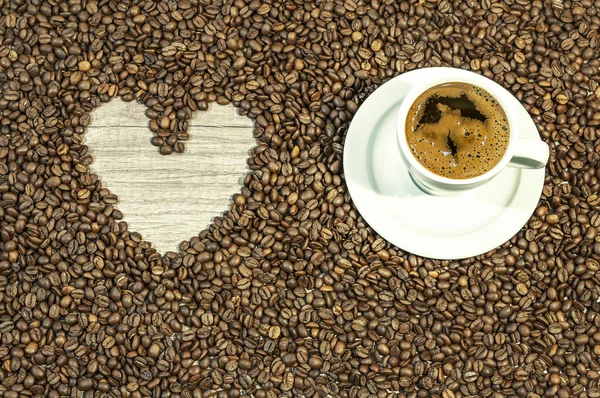 Fundo de grão de café com coração e xícara de café quente fresco na placa branca na mesa da cozinha — Fotografia de Stock