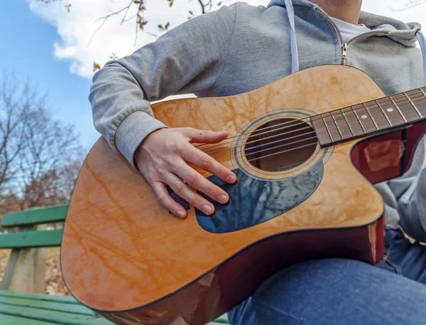 Young man sitting on bench in park and playing guitar — Stock Photo, Image