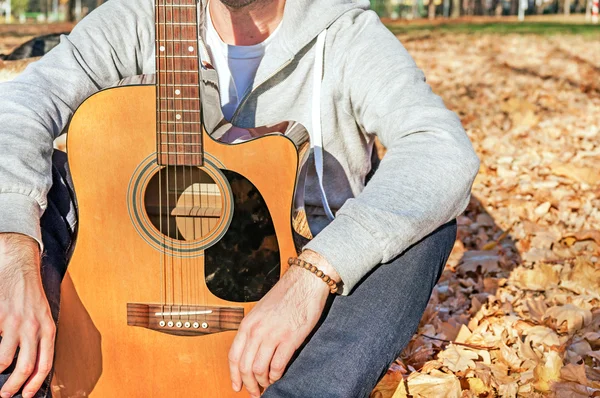 Young man sitting in autumn park on leaves and holding guitar — Stock Photo, Image