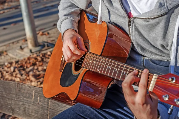 Young man sitting on wooden chair in park and playing guitar — Stock Photo, Image