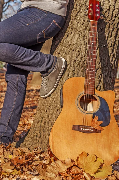 Man and guitar leaning on a tree in autumn park — Stock Photo, Image