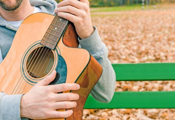 Young man holding acoustic guitar on bench in autumn park — Stock Photo, Image