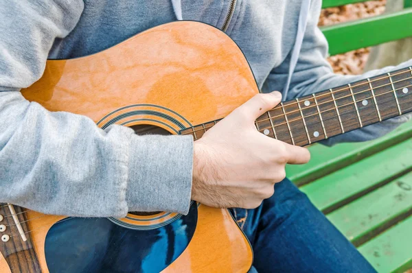 Young man holding acoustic guitar on bench in autumn park — Stock Photo, Image