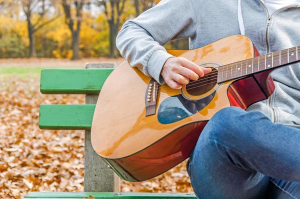 Young man playing acoustic guitar on bench in autumn park — Stock Photo, Image