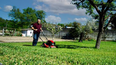 50-55 yaşlarında bir adam bahçedeki çimleri biçiyor.