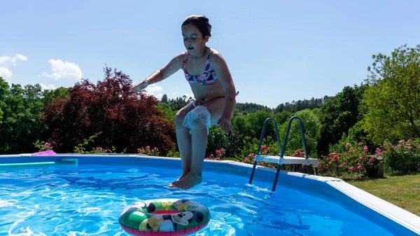 girl jumping into a pool standing upright and pointing towards a float