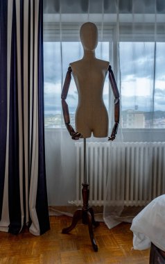 Bir dikiş mankeni ya da terziler, çizgili perdeleri olan büyük bir pencerenin yanında duran, eklemli ahşap kollarla büstü. Doğal ışıkla iç mekan ve arka planda kentsel manzara.