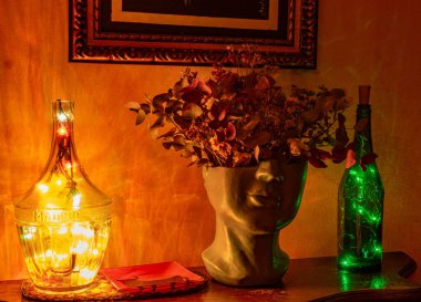 Dekoratif gece hayatı, biri berrak, biri yeşil olmak üzere iki cam şişe, LED ışıkları ve kurutulmuş yapraklarla dolu bir insan başlı vazo. Sıcak ve şenlikli bir atmosfer. Dekorasyon için ideal