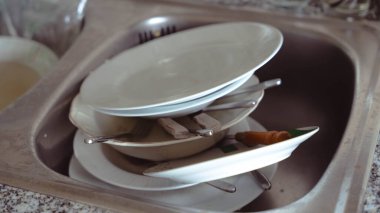 Kirli bulaşık, kase ve çatal bıçak yığını yıkanmayı bekleyen paslanmaz çelik bir lavabonun yakın çekimi. Görüntü bir yemeğin ya da ev işlerinin bittiğini gösteriyor..