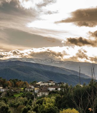 Etrafı yeşilliklerle çevrili küçük bir kırsal köyün panoramik manzarası, arkasında dramatik bulutlu bir gökyüzünün altında görkemli karla kaplı bir dağ. Doğanın ve kırsalın manzarası.