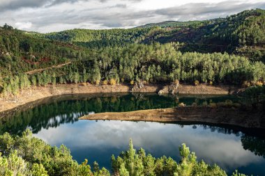 Bulutlu bir gökyüzünün altında yoğun bir çam ormanıyla çevrili dolambaçlı bir rezervuarın havadan görünüşü. Huzuru, uçsuz bucaksızlığı ve bozulmamış doğanın güzelliğini yansıtan bir görüntü..