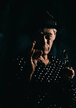 Şapkalı ve puantiyeli tişörtlü, el hareketi yapan yaşlı bir adamın sade sanatsal portresi. Karanlık, mistik bir arkaplan karşısında ifadeyi vurgulayan dramatik ışıklandırma.