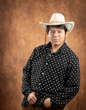 Açık renkli kovboy şapkası ve siyah noktalı siyah gömlek giyen bir adamın stüdyo portresi. Önden ciddi bir ifadeyle kahverengi arka plan ve sıcak ışıkta poz veriyor..