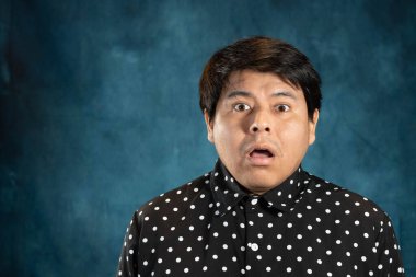 Aşırı şok veya şaşkınlık ifadesi olan bir adamın yakın çekim stüdyo portresi. Profesyonel ışıklandırmayla dokulu mavi arka planda siyah benekli bir gömlek giyiyor..