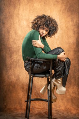 Taburede poz veren kıvırcık saçlı genç bir kadının stüdyo portresi. Kalıplı kahverengi bir arka planda yeşil bir süveter, deri pantolon ve platform botlar giyiyor..