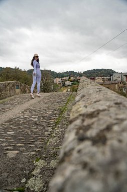 Eski bir taş köprüde yürüyen spor giysili genç bir kadının geniş açılı görüntüsü. Geleneksel mimari ve arka planda bulutlu gökyüzü ile kırsal ortam.