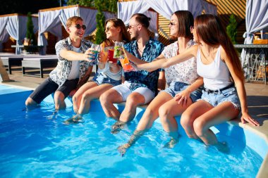 Bir grup arkadaş havuz başında eğleniyor bardakları tokuşturup taze kokteyllerle güneşli yaz gününde havuzda oturuyorlar. İnsanlar tropikal tatillerde lüks villada içki içiyorlar..